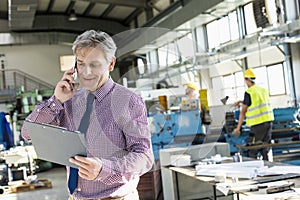 Mature male supervisor looking at clipboard while talking on mobile phone in industry