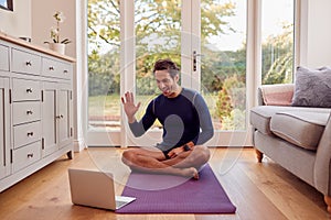 Mature Male Teacher Sitting On Mat At Home With Laptop Giving Online Yoga Class