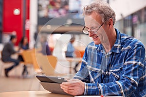 Mature Male Teacher Or Student With Digital Tablet Working At Table In College Hall