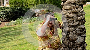 Mature man in aloha shirt looks through binoculars closed to palm trunk in park