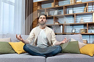 Mature man alone at home resting after work sitting in lotus position on sofa in living room meditating and relaxing