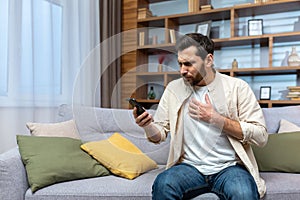 Mature man with beard alone at home reading bad news online on phone while sitting on sofa in living room at home