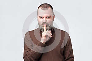 Mature man with beard in brown shirt making silence gesture