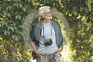 A mature man with a beard walks in the park with a camera. Active pastime in old age