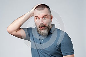 Mature man in blue t-shirt looks with frightened expression as sees something awful in front