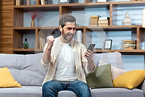 Mature man celebrating victory and successful achievement, got news online message on app using phone sitting on sofa in