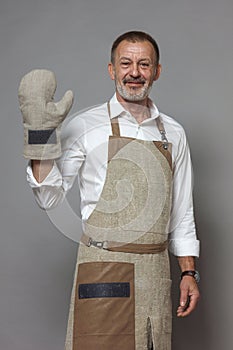 Mature man in a chefs apron waves, greets with an oven mitt and smiles.