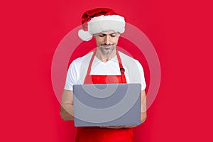 mature man in christmas santa hat and red apron hold laptop