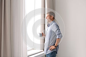 Mature man with cup of drink near window with open curtains at home