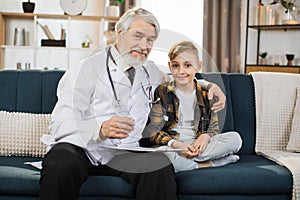 Mature man doctor in white coat and stethoscope on shoulders talking to sick boy patient