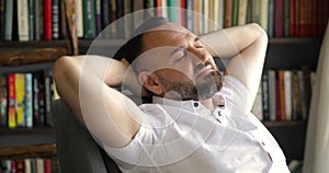 Mature man is dreaming snoozing in armchair in library with hands behind head.