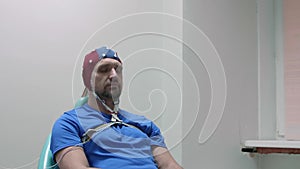 Mature man in EEG cap sits on chair with eyes closed.