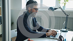 Mature man in elegant suit putting on glasses and working with laptop in office