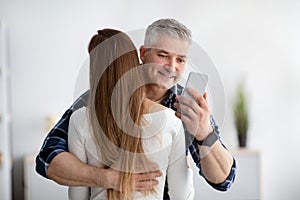 Mature man embracing his wife, stuck in smartphone, not paying attention, texting his lover, going through family crisis