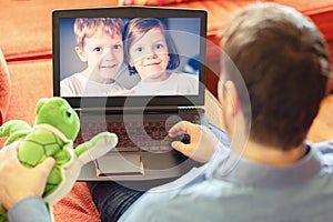 Mature man father lying on sofa and communicate trough video call on laptop with his kids, a little boy and girl showing them toys photo