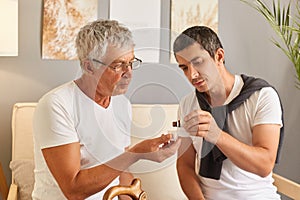 Mature man feeling unwell his adult son giving medicine for sick father putting drops into glasses helping mature male to feel