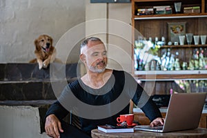 Mature man freelancer working with laptop and phone holding red cup of coffee and brown dog on background