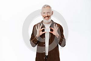 Mature man with grey hair smiling, showing credit card, senior guy recommending bank, standing over white background