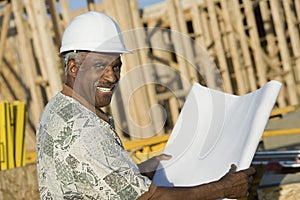 Mature Man In Hardhat With Blueprint At House Construction Site