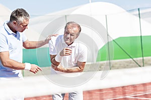 Mature man helping senior friend with elbow injury while playing tennis on sunny day