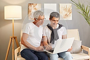 Mature man with his adult son wearing casual clothing using portable computer together young guy teaching to use laptop browsing