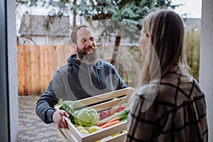 Mature man holding crate with vegetales and fruit and delivering it to woman standing at doorway. photo