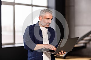 Mature man holding using laptop standing leaning on white wall, posing looking at camera