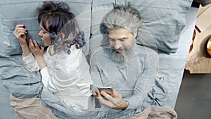 Mature man husband using smartphone texting while young woman sleeping in bed