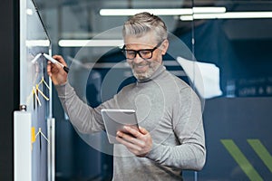 Mature man inside office near whiteboard with colored notes writing down strategy and business plan, gray haired