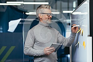 Mature man inside office near whiteboard with colored notes writing down strategy and business plan, gray haired