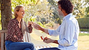 Mature Man Kneeling And Proposing To Surprised Woman Sitting In Park With Engagement Ring In Box