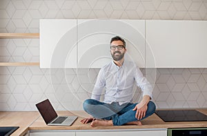 A mature man with laptop sitting in kitchen in unfurnished new house.