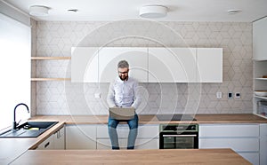 A mature man with laptop sitting in kitchen in unfurnished new house.