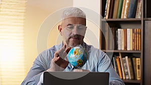 Mature man looking at globe map of world and dreaming of vacation, sitting near laptop at home