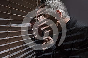 Mature man looking out of a window with blinds casting shadows