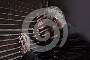 Mature man looking out of a window with blinds casting shadows