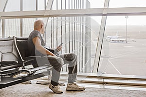 A mature man looks through a panoramic glass wall at the airfield, sitting in the departure hall of the airport