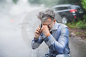 Mature man making a phone call after a car accident, smoke in the background.