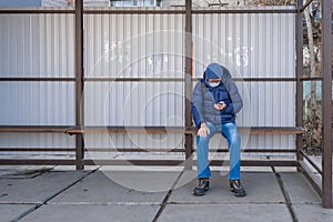 A mature man in a medical mask sits at a public transport stop in autumn and waits for a bus  tram  trolleybus