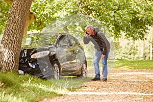 Mature Man Next To Car Crashed Into Tree Inspecting Accident Damage And Calling Emergency Services