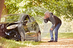 Mature Man Next To Car Crashed Into Tree Inspecting Accident Damage And Calling Emergency Services