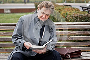 A mature man in an old gray cloak sits on a bench and enthusiastically reads a thick black book. Salesman, member of a religious