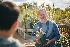 Mature Man Outdoors In Garden Centre Asking Advice From Sales Assistant Choosing And Buying Rose