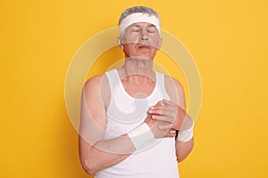 Mature man posing against yellow wall with closed eyes and touching his chest, feels pain in heart, needs treatment, has heart