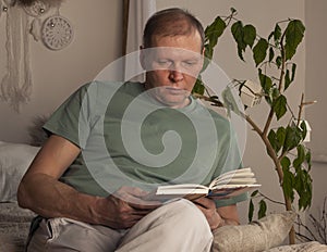 Mature man reading book in modern trendy cozy interior with plant in evening, alone