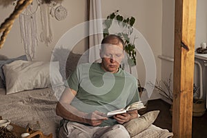 Mature man reading book in modern trendy cozy interior with plant in evening, alone
