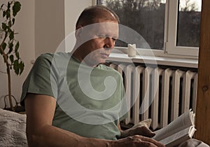 Mature man reading book in modern trendy cozy interior with plant in evening, alone