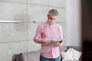 Mature man reading document while standing against wall at home