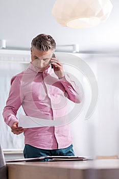 Mature man reading document while talking on smart phone at table in apartment