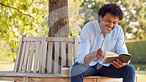 Mature Man Relaxing Sitting On Park Bench Under Tree Reading Book With Takeaway Coffee